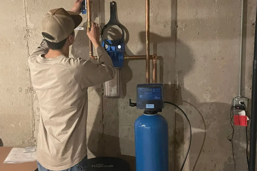 A person installing a water softener system with copper pipes in a basement against a concrete wall.
